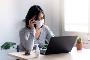 Young Quarantined Woman Talking On The Phone Wearing Medical Face Mask And With Hand Sanitizer Working From Home Office Due To Corona Virus Outbreak