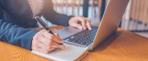 Business Women Hand Are Taking Notes On Paper With A Black Pen, And She Is Using A Laptop Computer On A Wooden Desk In The Office.Web Banner.