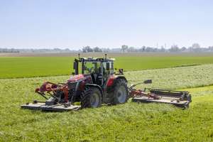 A red tractor with large wheels mowing a lush green field under a clear blue sky, with wind turbines visible in the distant background.