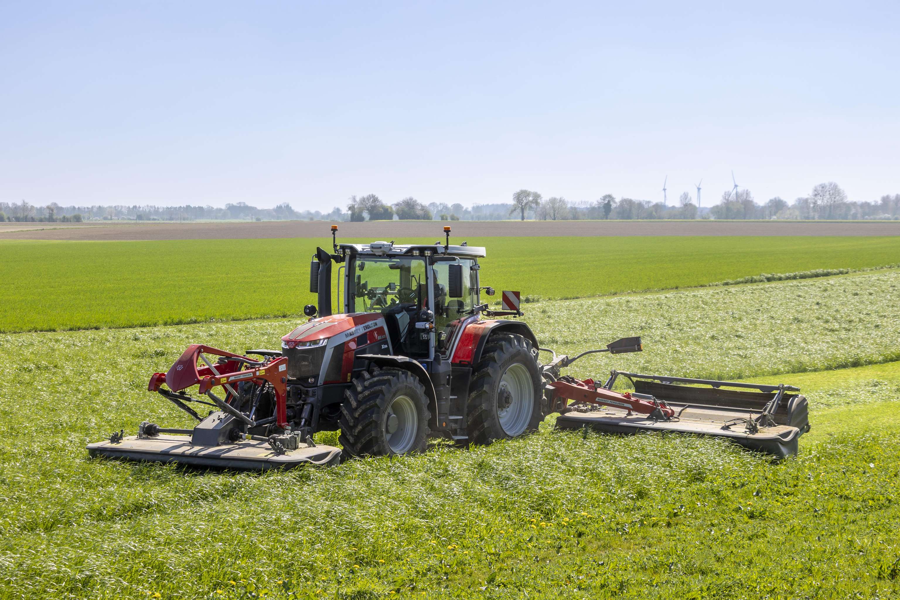 A red tractor with large wheels mowing a lush green field under a clear blue sky, with wind turbines visible in the distant background.