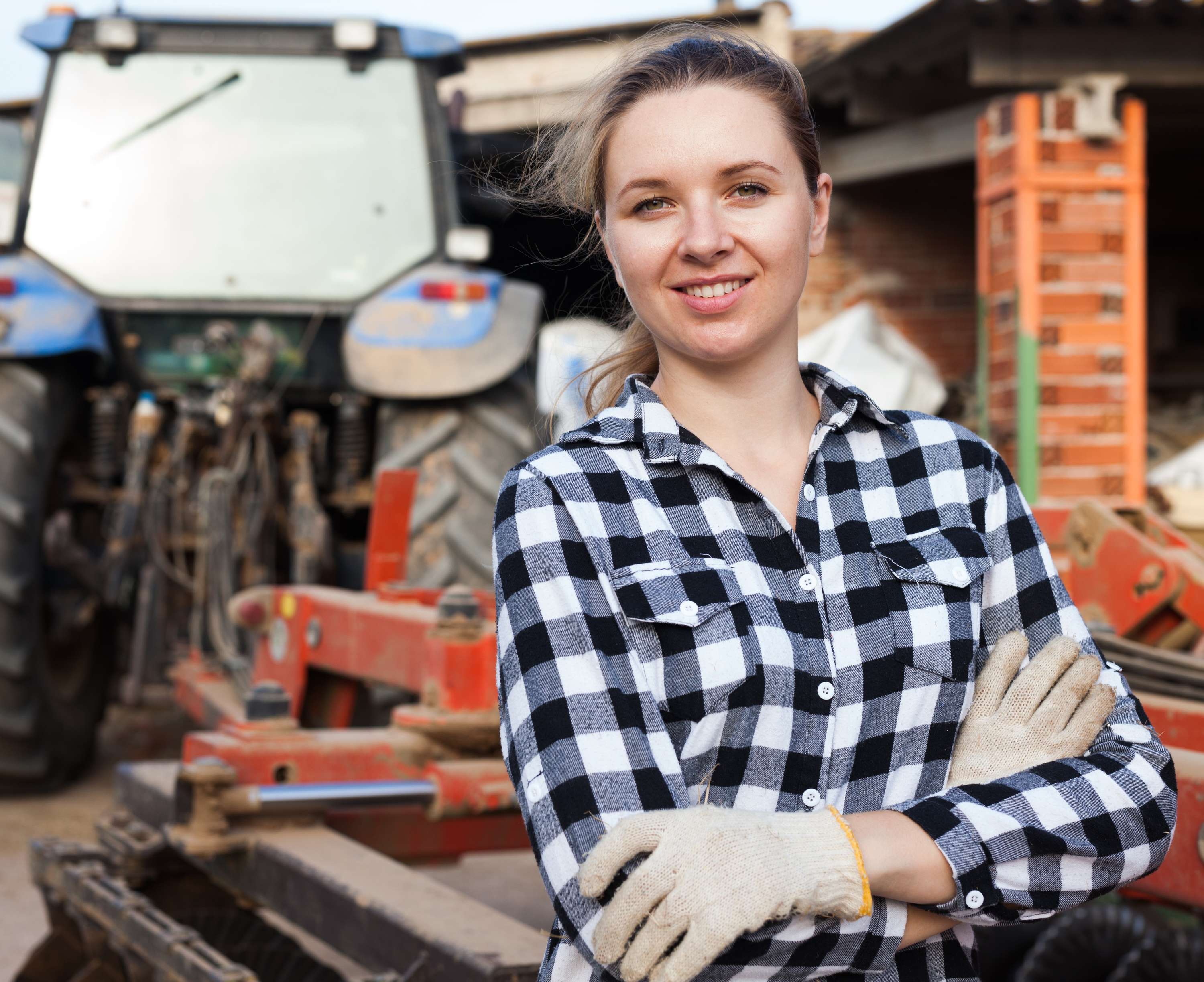 Junge Bäuerin im karierten Hemd mit Arbeitshandschuhen steht lächelnd vor einem blauen Traktor auf einem Bauernhof.