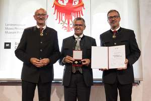 Three men in formal attire stand smiling; the center man holds a medal, and the right man displays a certificate. A logo is in the background.
