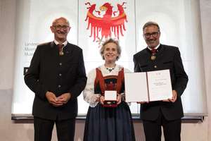 Three people in traditional attire stand smiling, holding an award and certificate, with a red emblem in the background.