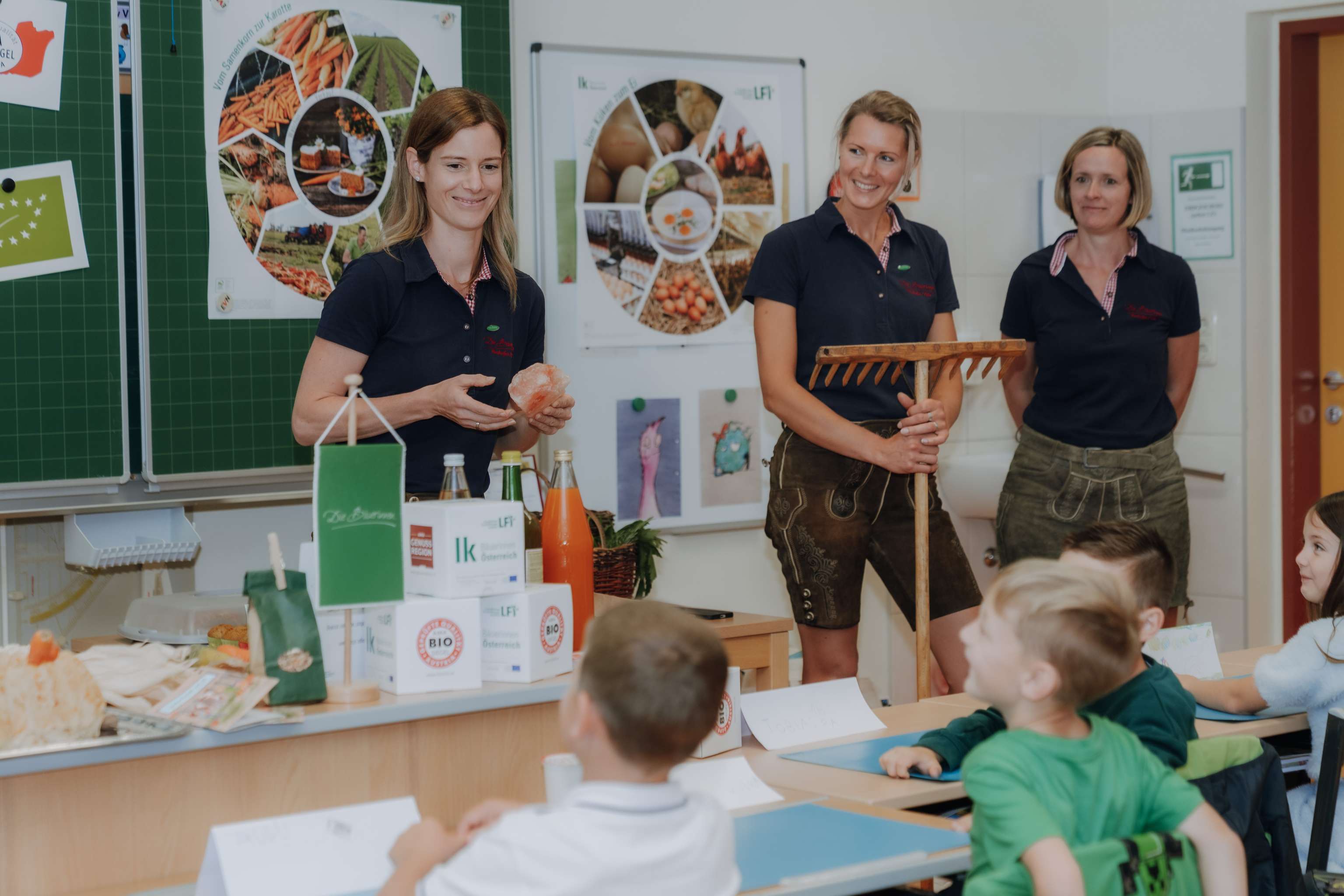 Drei Frauen in dunklen Poloshirts präsentieren Kindern im Klassenzimmer landwirtschaftliche Produkte und Bio-Lebensmittel mit Lehrpostern im Hintergrund.