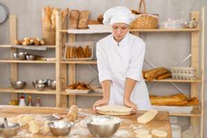 Junge Bäckerin in weißer Uniform knetet Teig auf einem bemehlten Holztisch in einer Bäckerei mit Brotregalen im Hintergrund.