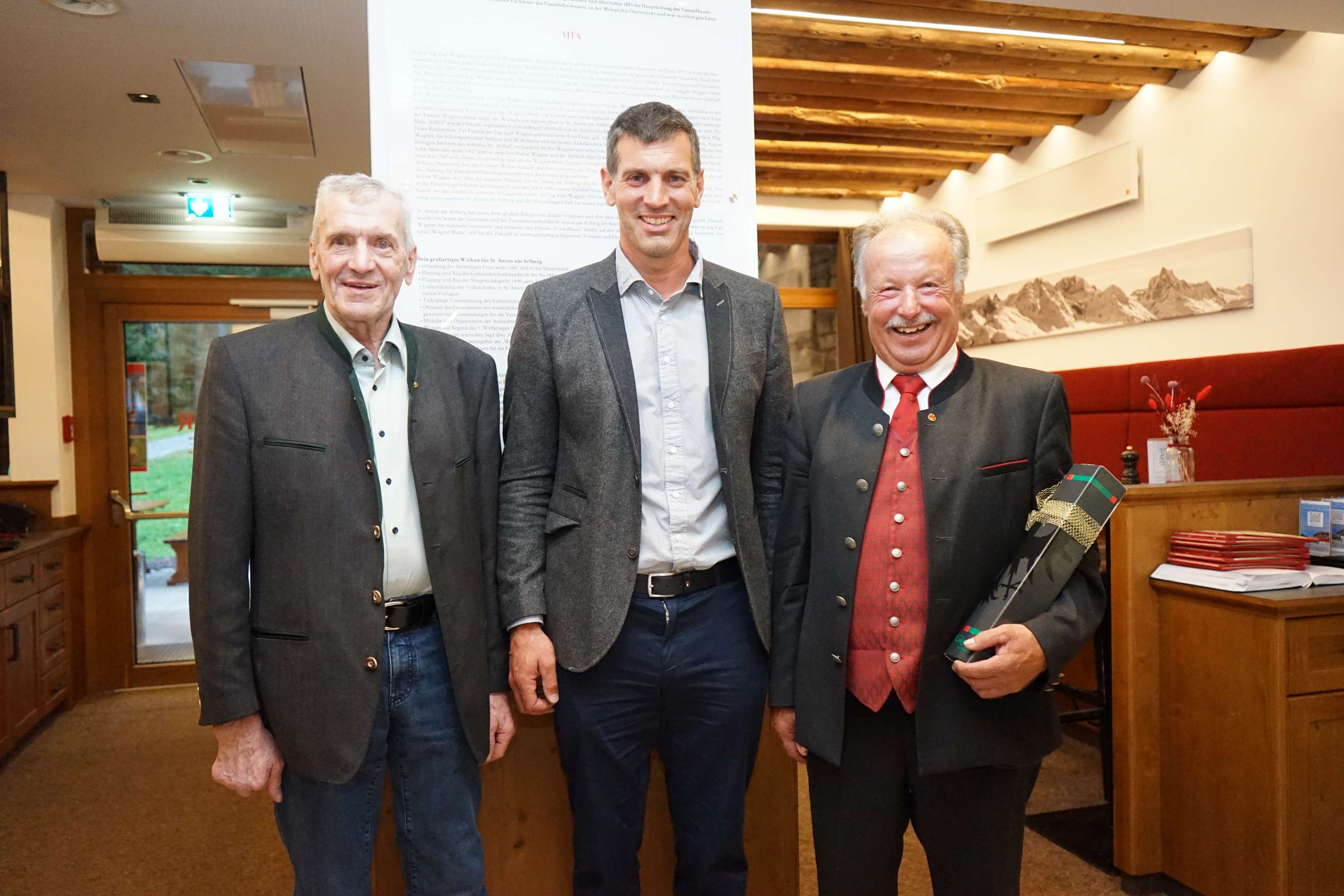 Drei Männer in Trachtenkleidung posieren lächelnd bei einer Veranstaltung, einer hält ein Geschenk in der Hand.