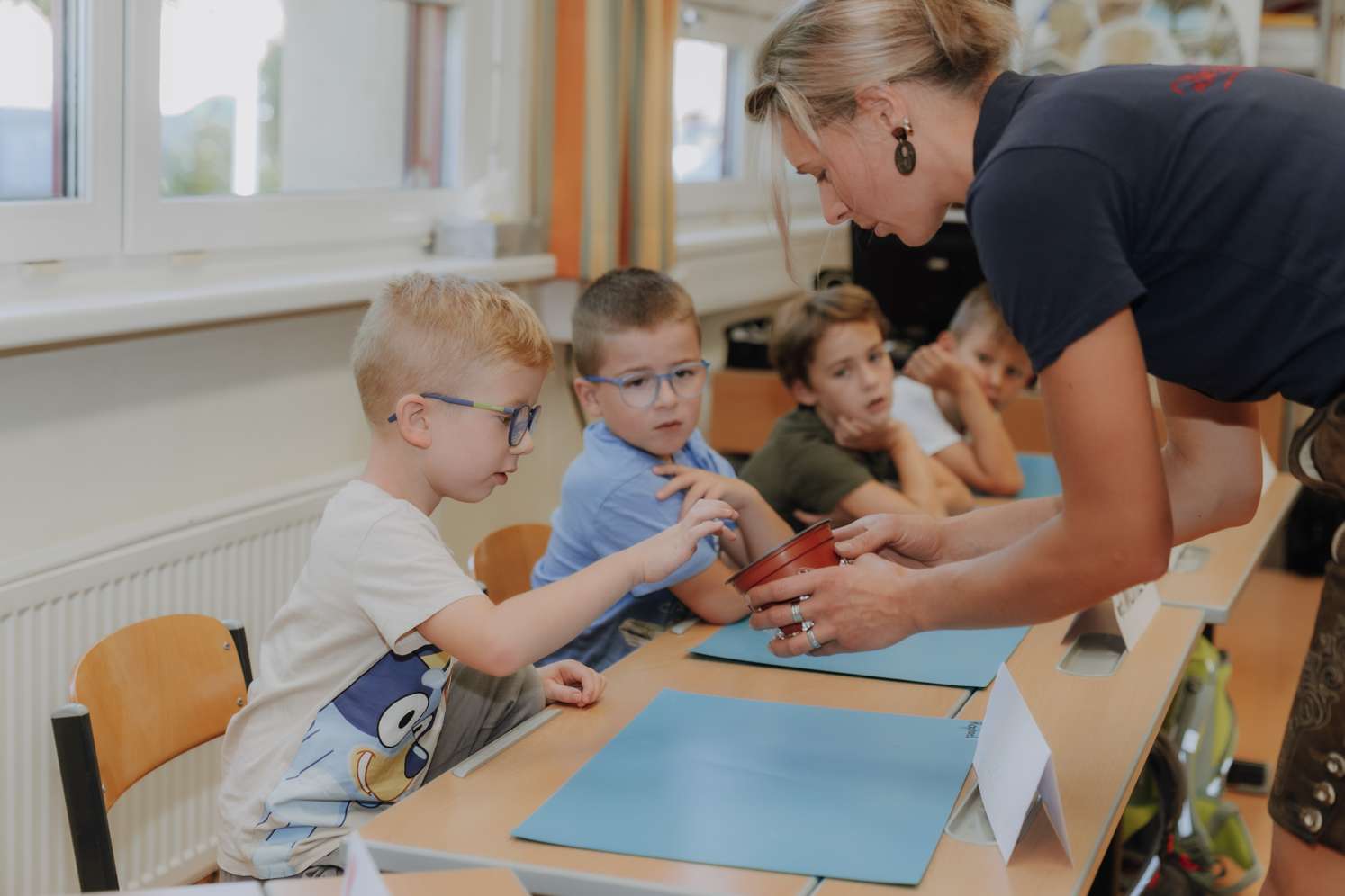 Lehrerin reicht einem kleinen Jungen mit Brille einen Blumentopf, während weitere Kinder am Tisch im Klassenzimmer zuschauen.