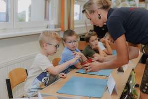 Lehrerin reicht einem kleinen Jungen mit Brille einen Blumentopf, während weitere Kinder am Tisch im Klassenzimmer zuschauen.