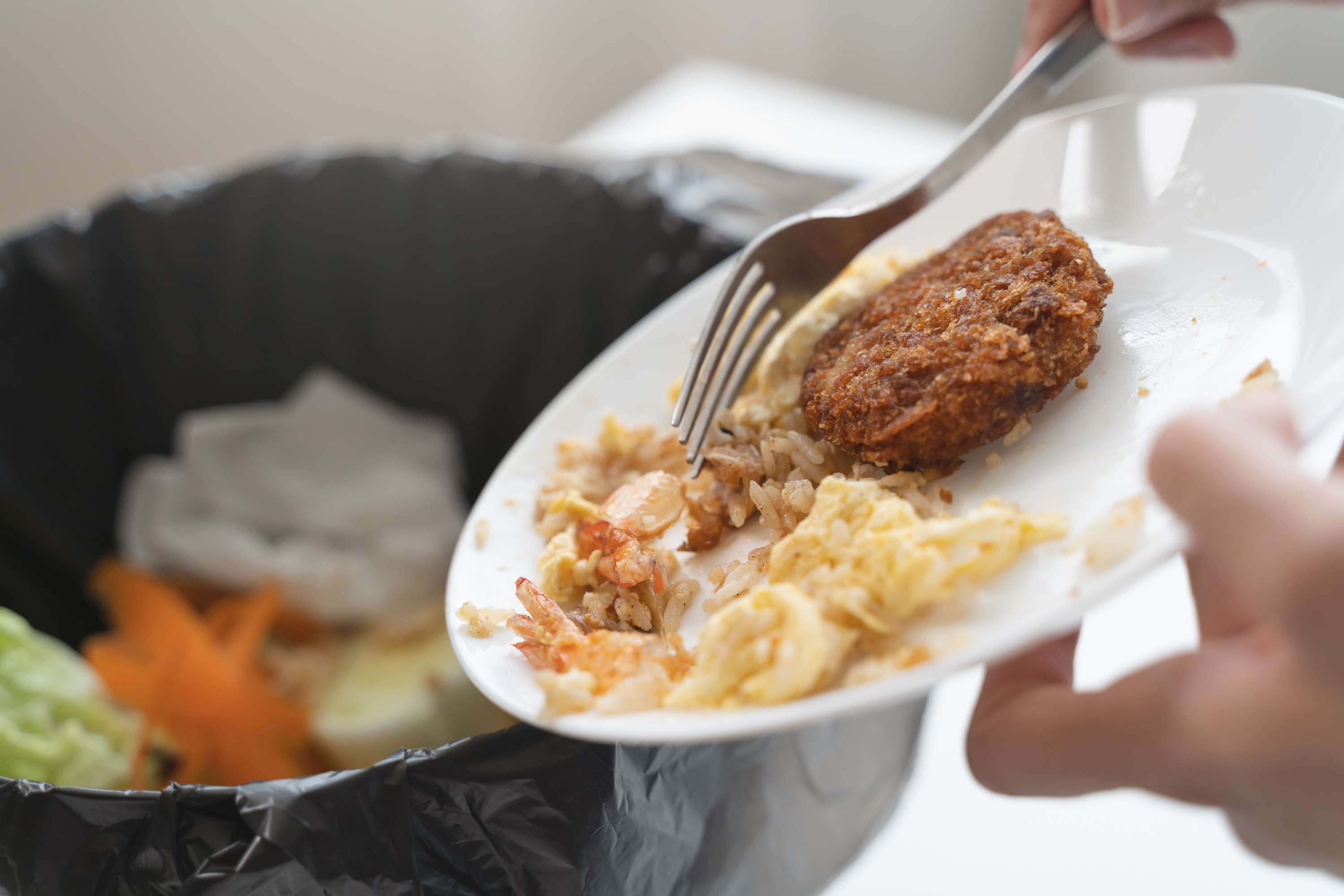A person scraping leftover food from a plate into a trash bin, including rice, vegetables, and a breaded item, with a fork in hand.