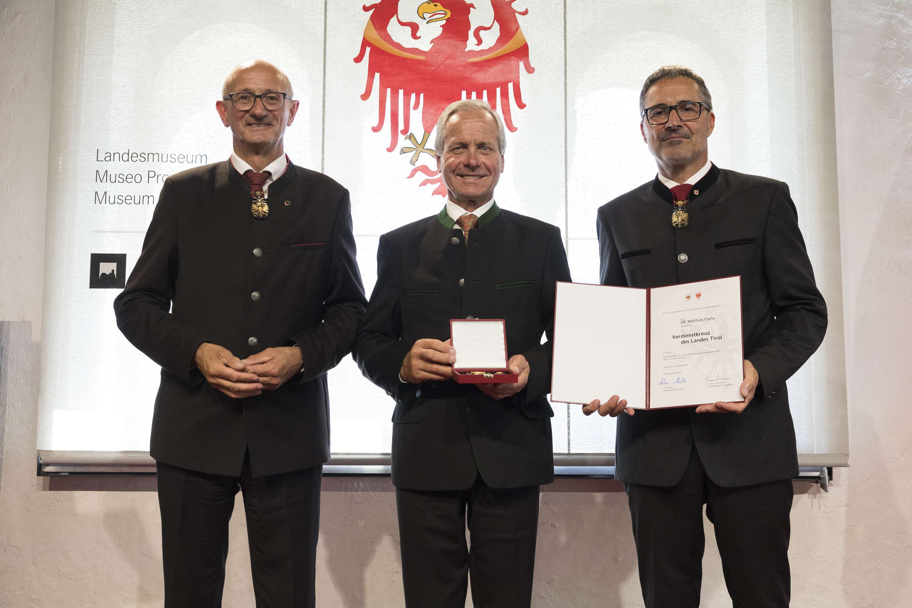 Three men in traditional attire stand proudly; the middle one holds a medal, and the man on the right displays a certificate.