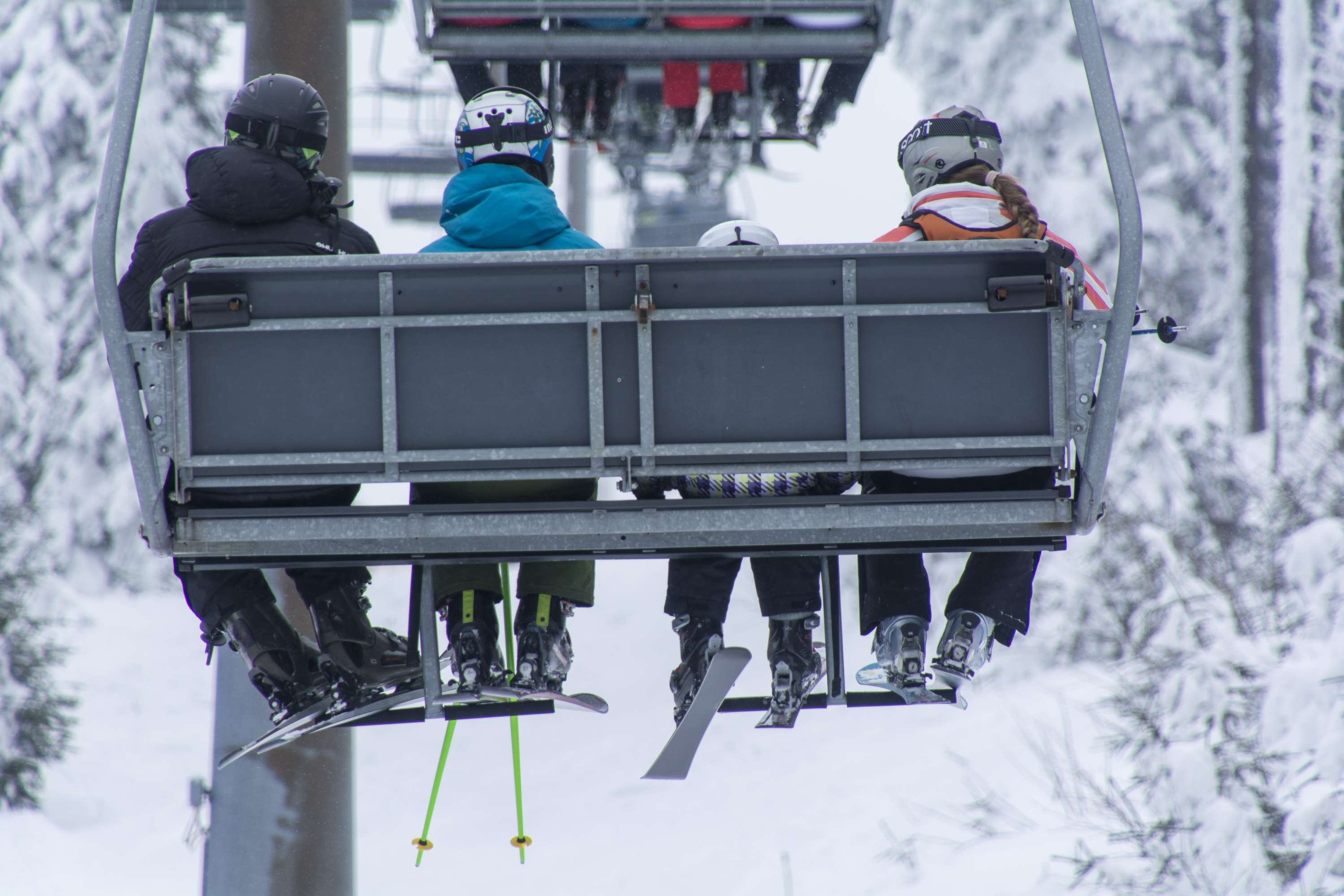 Vier Personen mit Skiern und Snowboards auf einem Sessellift in schneebedeckter Umgebung, von hinten gesehen.