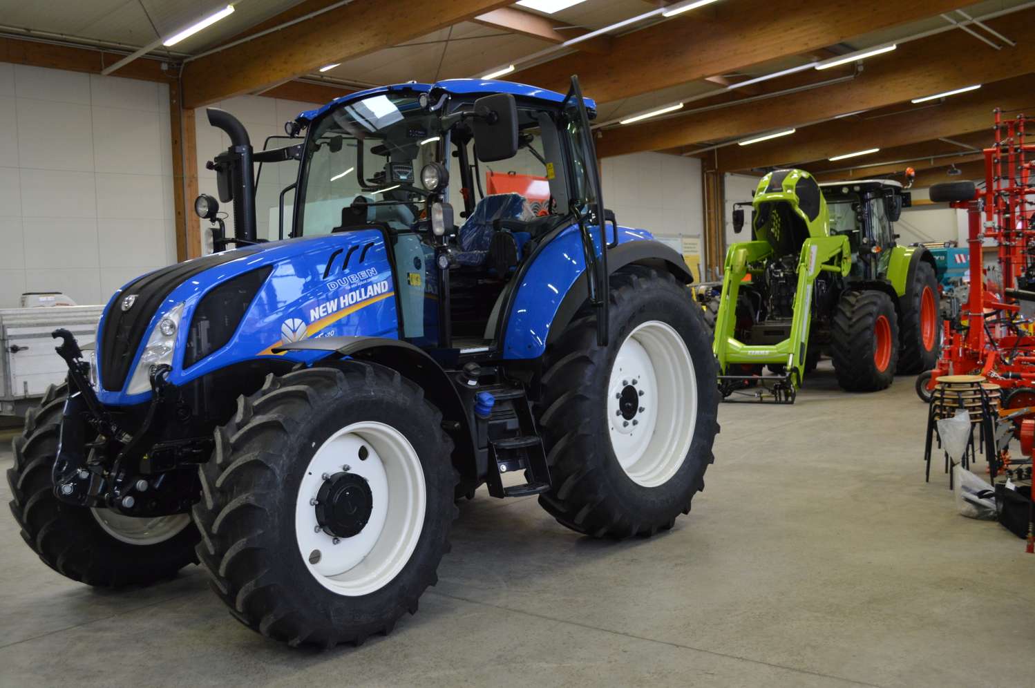 Blauer New Holland Traktor in einer Ausstellungshalle, im Hintergrund ein grüner Claas Traktor und rote Landmaschinen.