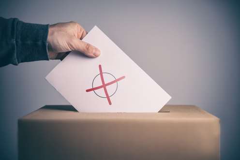 Eine Hand legt einen Wahlschein mit einem roten Kreuz in eine braune Wahlurne.