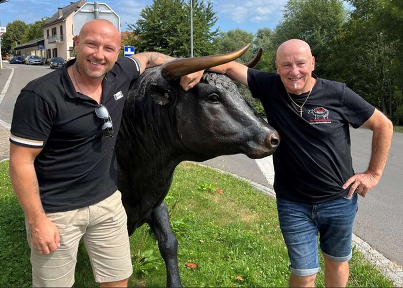 Zwei Männer in schwarzen T-Shirts posieren lächelnd neben einer schwarzen Stierstatue mit goldenen Hörnern auf einer Grünfläche.