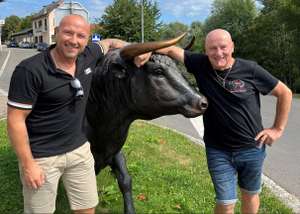 Zwei Männer in schwarzen T-Shirts posieren lächelnd neben einer schwarzen Stierstatue mit goldenen Hörnern auf einer Grünfläche.