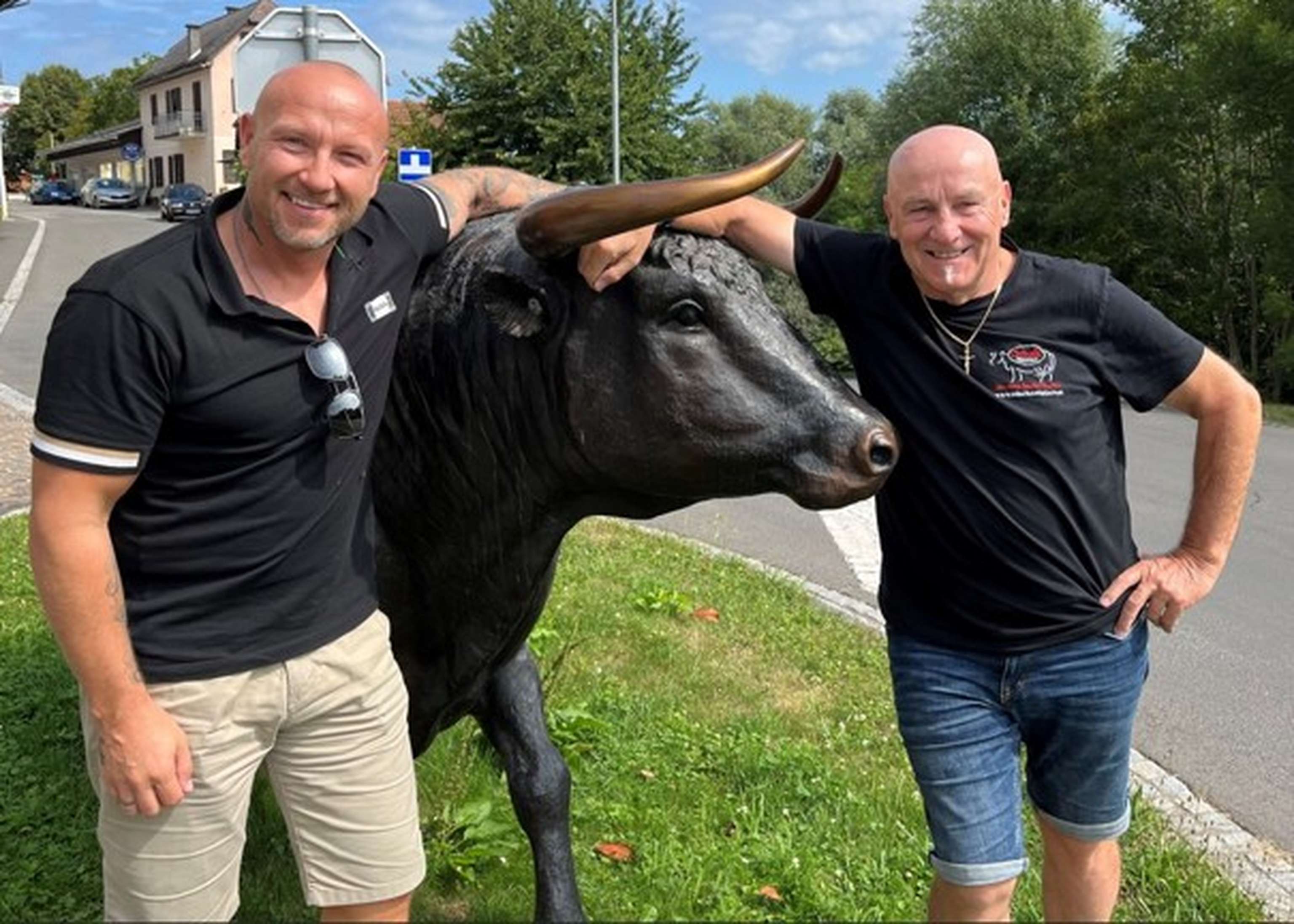Zwei Männer in schwarzen T-Shirts posieren lächelnd neben einer schwarzen Stierstatue mit goldenen Hörnern auf einer Grünfläche.