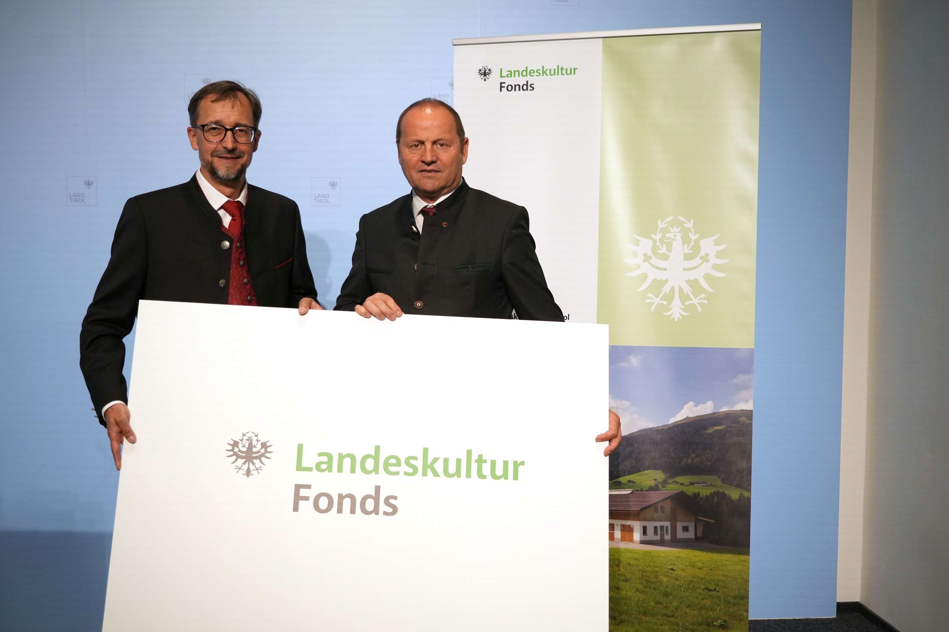 Zwei Männer in traditioneller Kleidung halten ein großes Schild mit der Aufschrift "Landeskultur Fonds" vor einem Werbebanner.