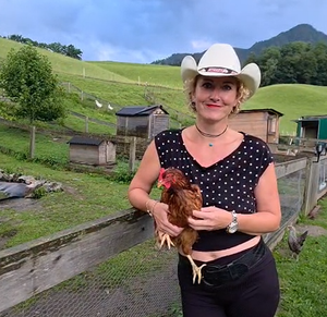 Frau mit weißem Cowboyhut hält ein braunes Huhn auf einem Bauernhof mit grünen Hügeln im Hintergrund.
