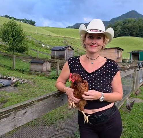 Frau mit weißem Cowboyhut hält ein braunes Huhn auf einem Bauernhof mit grünen Hügeln im Hintergrund.