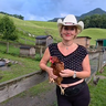 Frau mit weißem Cowboyhut hält ein braunes Huhn auf einem Bauernhof mit grünen Hügeln im Hintergrund.