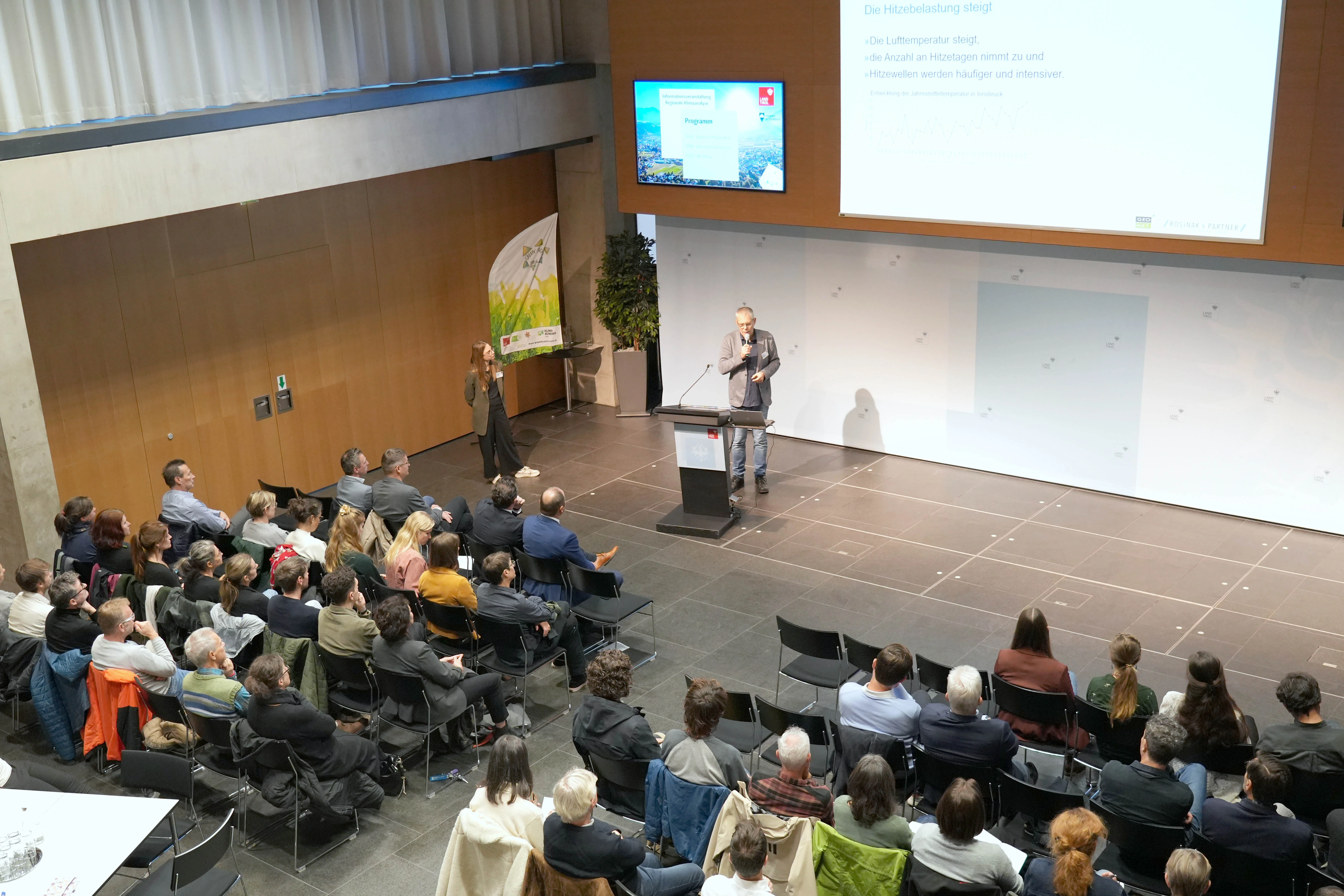 A speaker presents to a seated audience in a modern conference hall, with a large screen displaying a presentation.