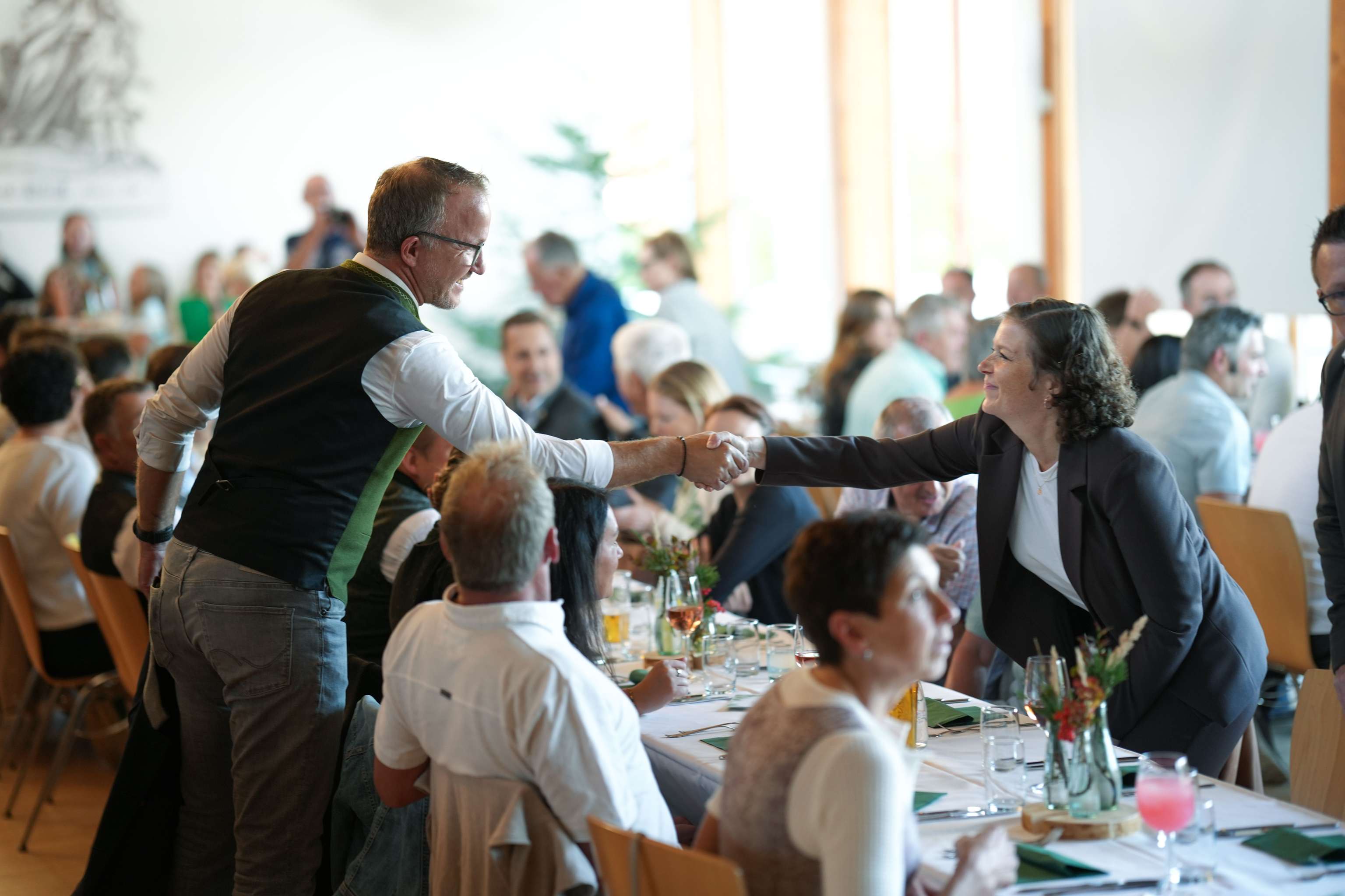 Landesrat Gantner reicht einem Festgast die Hand bei einer festlichen Veranstaltung in einem voll besetzten Saal mit gedeckten Tischen.