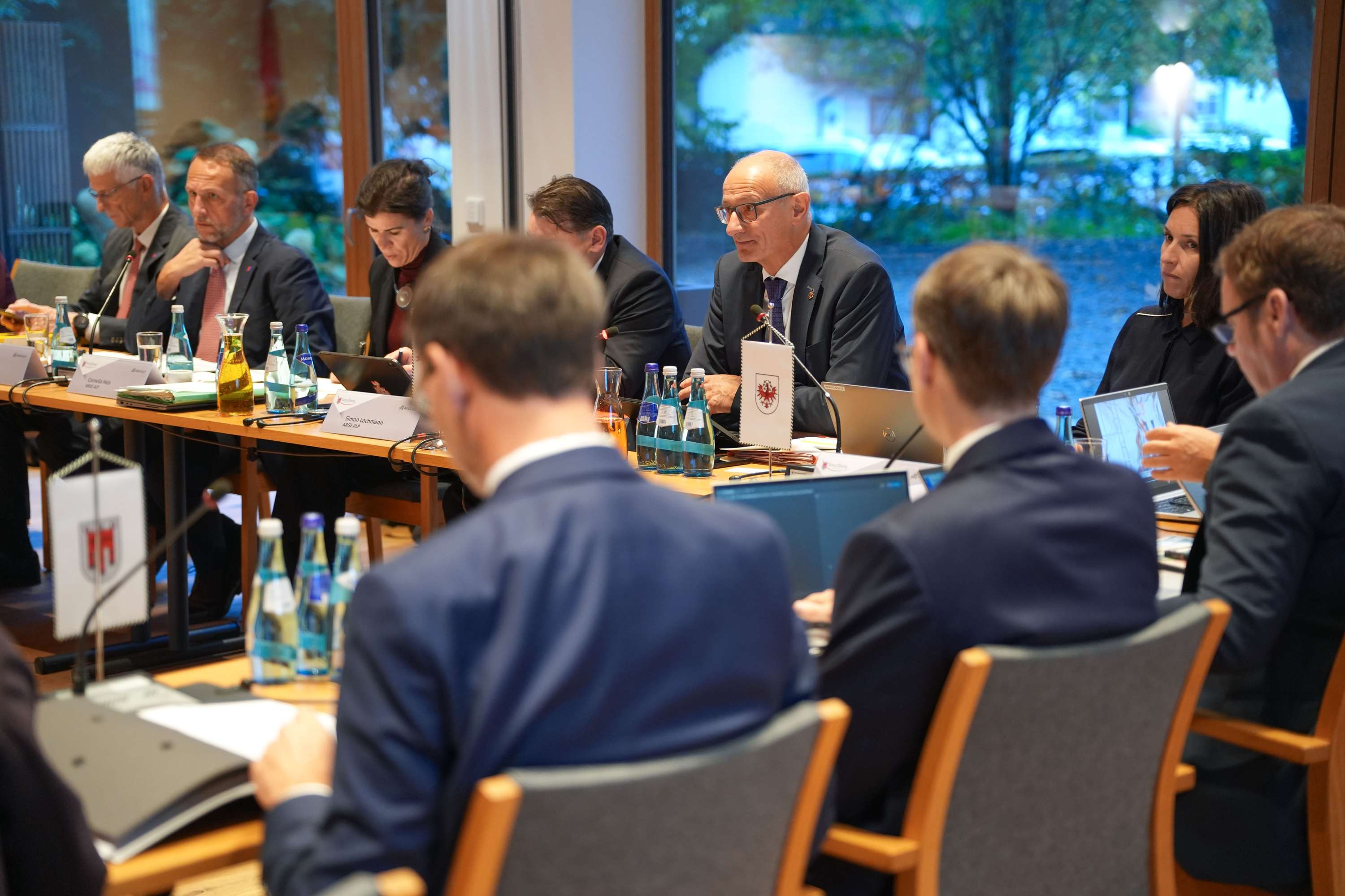 Politische Sitzung mit mehreren Personen in Anzügen an einem Konferenztisch mit Namensschildern, Mikrofonen und Wasserflaschen.