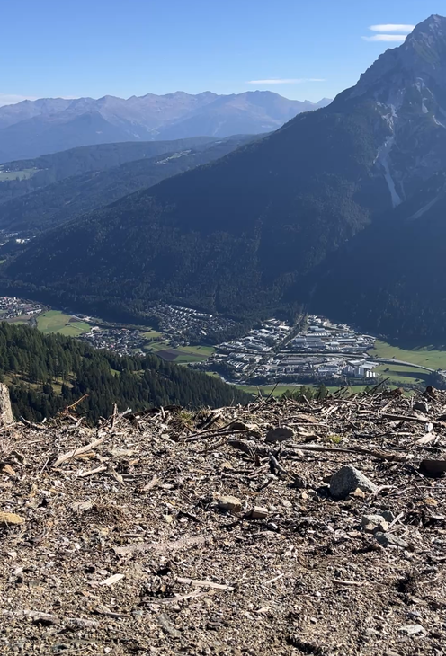 Blick vom steinigen Berghang auf ein Alpental mit Ortschaft, umgeben von bewaldeten Bergen und blauem Himmel.