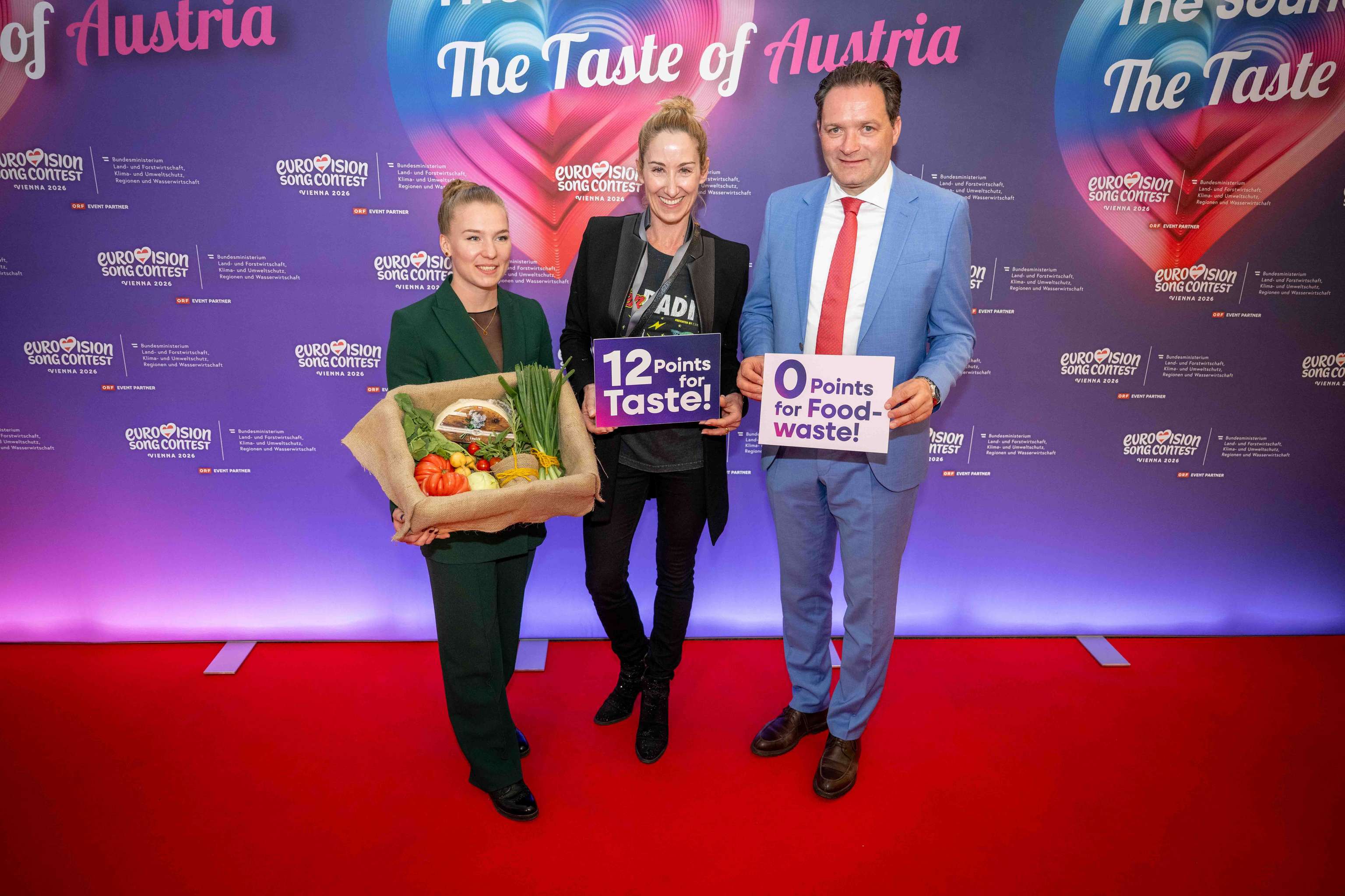 Drei Personen auf rotem Teppich, halten Schilder mit "12 Points for Taste" und "0 Points for Food-waste" mit Gemüsekorb. Hintergrund: Eurovision-Logo.
