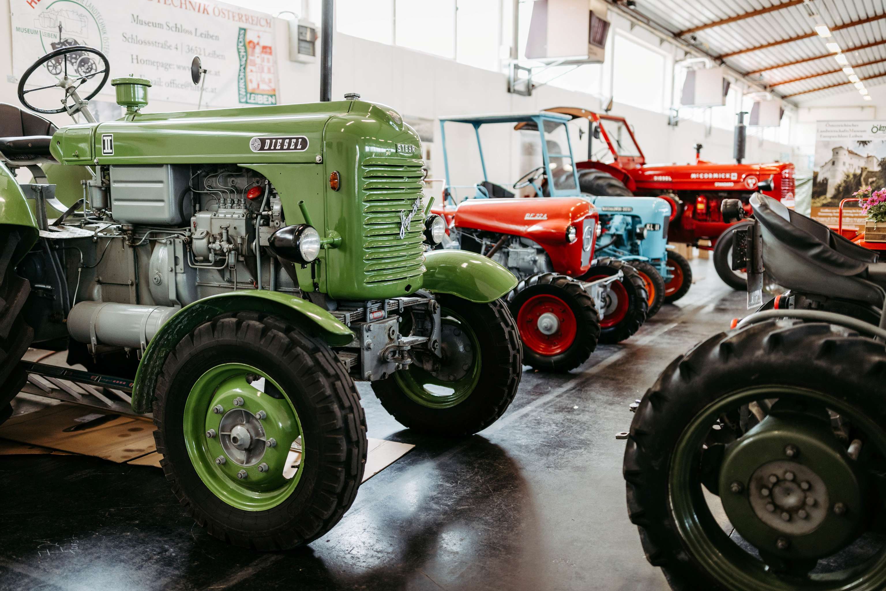Reihe alter Traktoren in verschiedenen Farben in einer Ausstellungshalle, mit einem grünen Traktor im Vordergrund.