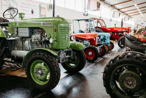 Reihe alter Traktoren in verschiedenen Farben in einer Ausstellungshalle, mit einem grünen Traktor im Vordergrund.