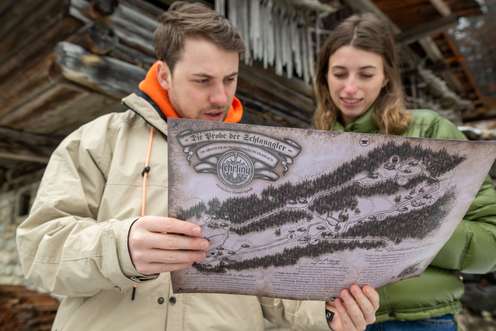 Ein Mann und eine Frau betrachten eine Karte im Freien vor einem Holzgebäude. Sie wirken konzentriert und neugierig.