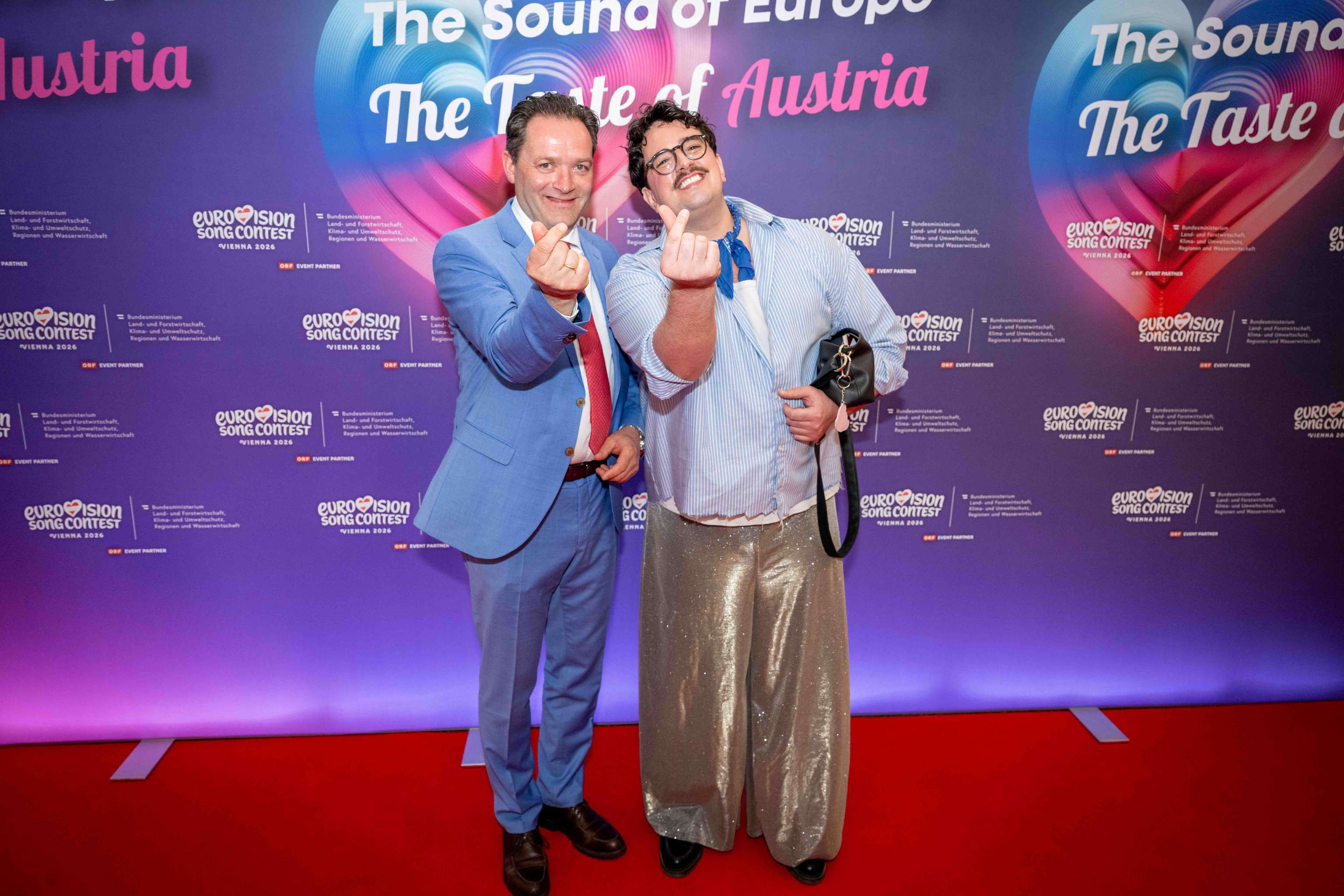 Zwei Männer posieren auf dem roten Teppich beim Eurovision Event, einer in blauem Anzug, der andere in glitzernder Hose.