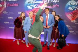 Gruppe von Menschen posiert für ein Selfie auf einem roten Teppich vor einem Plakat für den Eurovision Song Contest in Wien 2026.