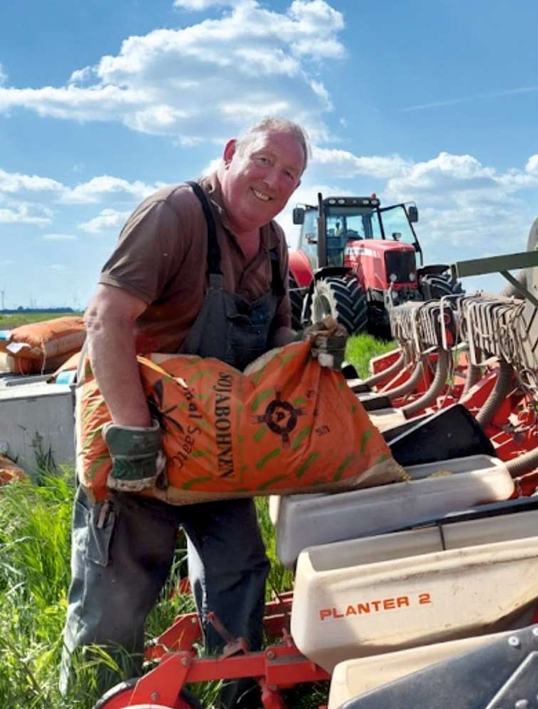 Smiling man in overalls and gloves holds a bag of seeds beside farming equipment, with a red tractor in the background under a blue sky.