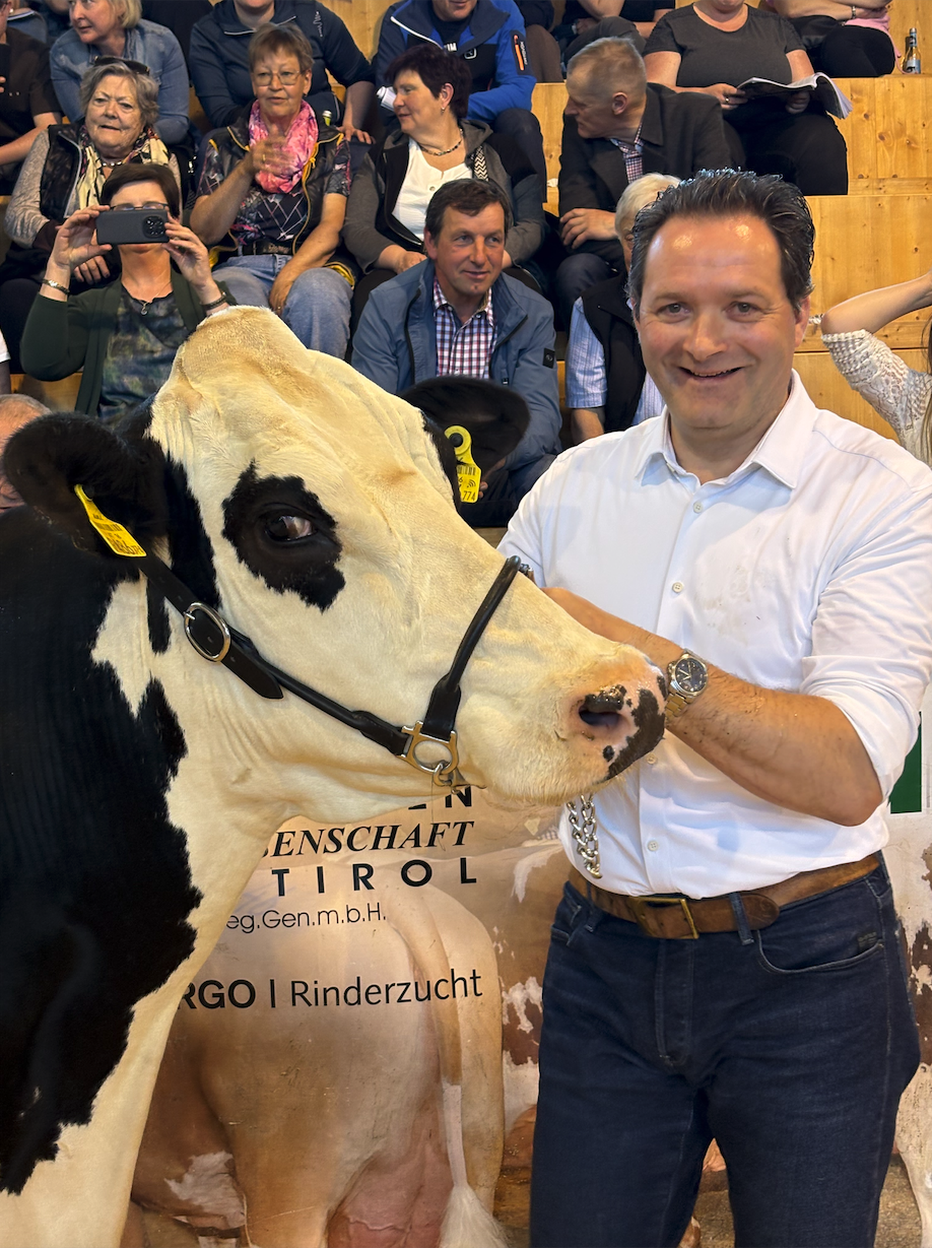 Ein Mann neben einer schwarz-weißen Kuh in einer Halle mit Zuschauern im Hintergrund.