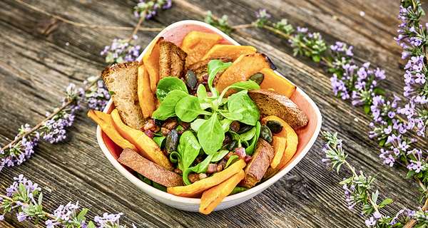 Feldsalat mit geröstetem Kürbis, Speck, Kernen und Croutons in weißer Schale auf Holztisch mit lila Blumen dekoriert.