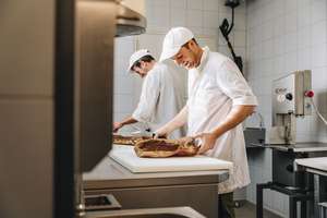 Zwei Köche in weißer Arbeitskleidung schneiden Fleisch in einer gewerblichen Küche. Einer lächelt, während sie konzentriert arbeiten.