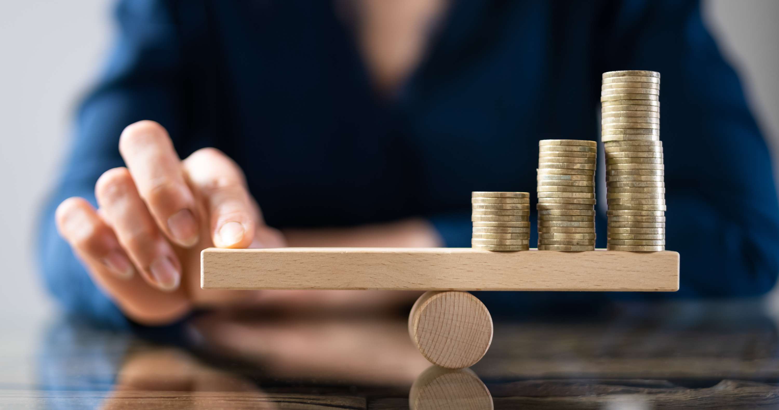 Hand balancing a wooden seesaw topped with three stacks of coins of increasing height, symbolizing financial growth.