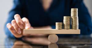 Hand balancing a wooden seesaw topped with three stacks of coins of increasing height, symbolizing financial growth.