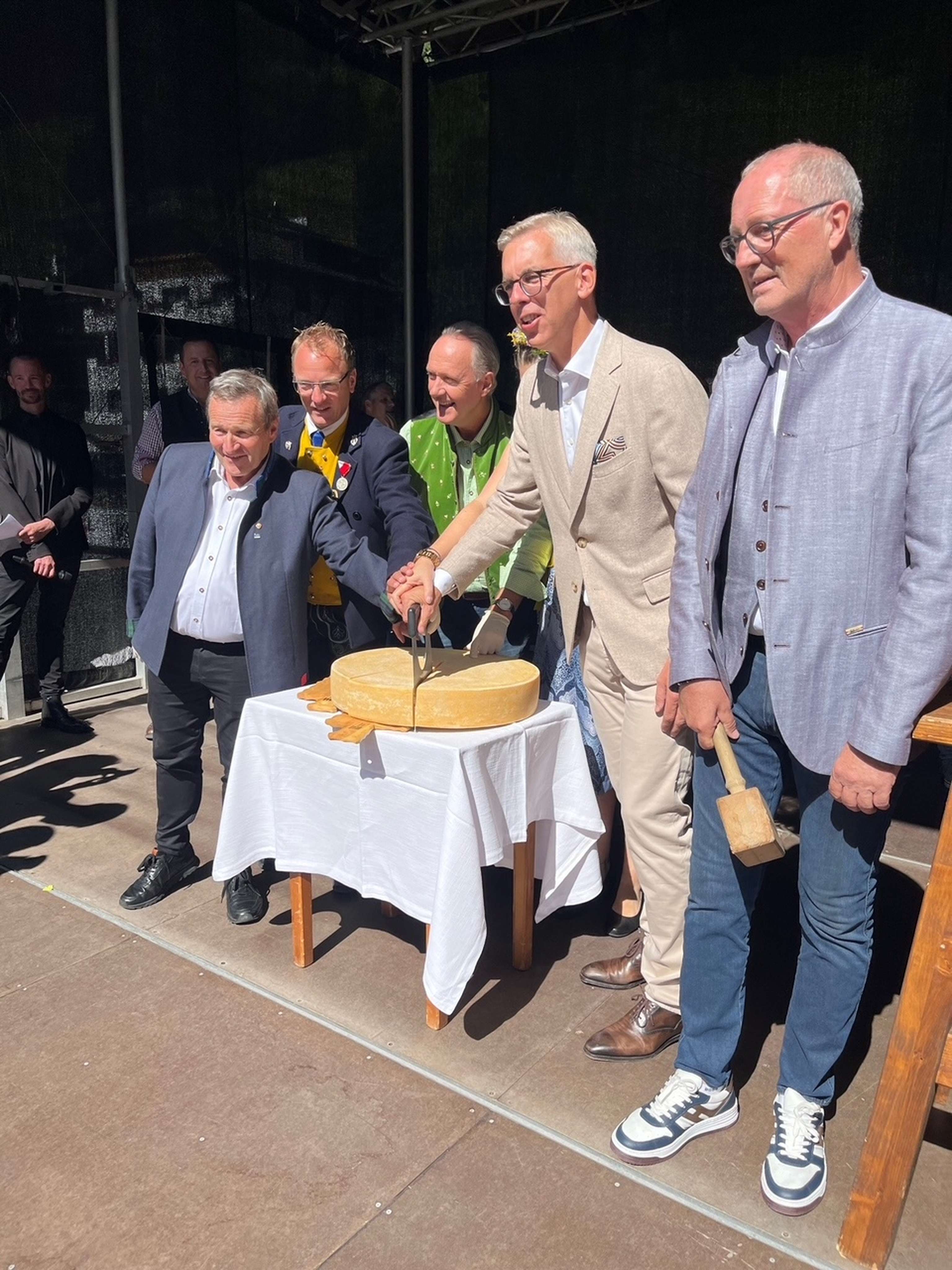 Mehrere Männer in festlicher Kleidung schneiden gemeinsam einen großen Käselaib auf einem weißen Tisch beim Alkmäsemarkttag in Ischgl..