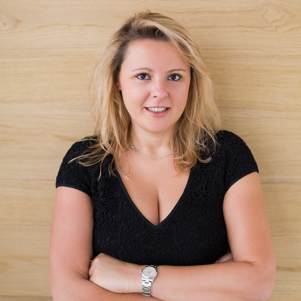 Woman with blonde hair in a black top, arms crossed, smiling in front of a wooden background.