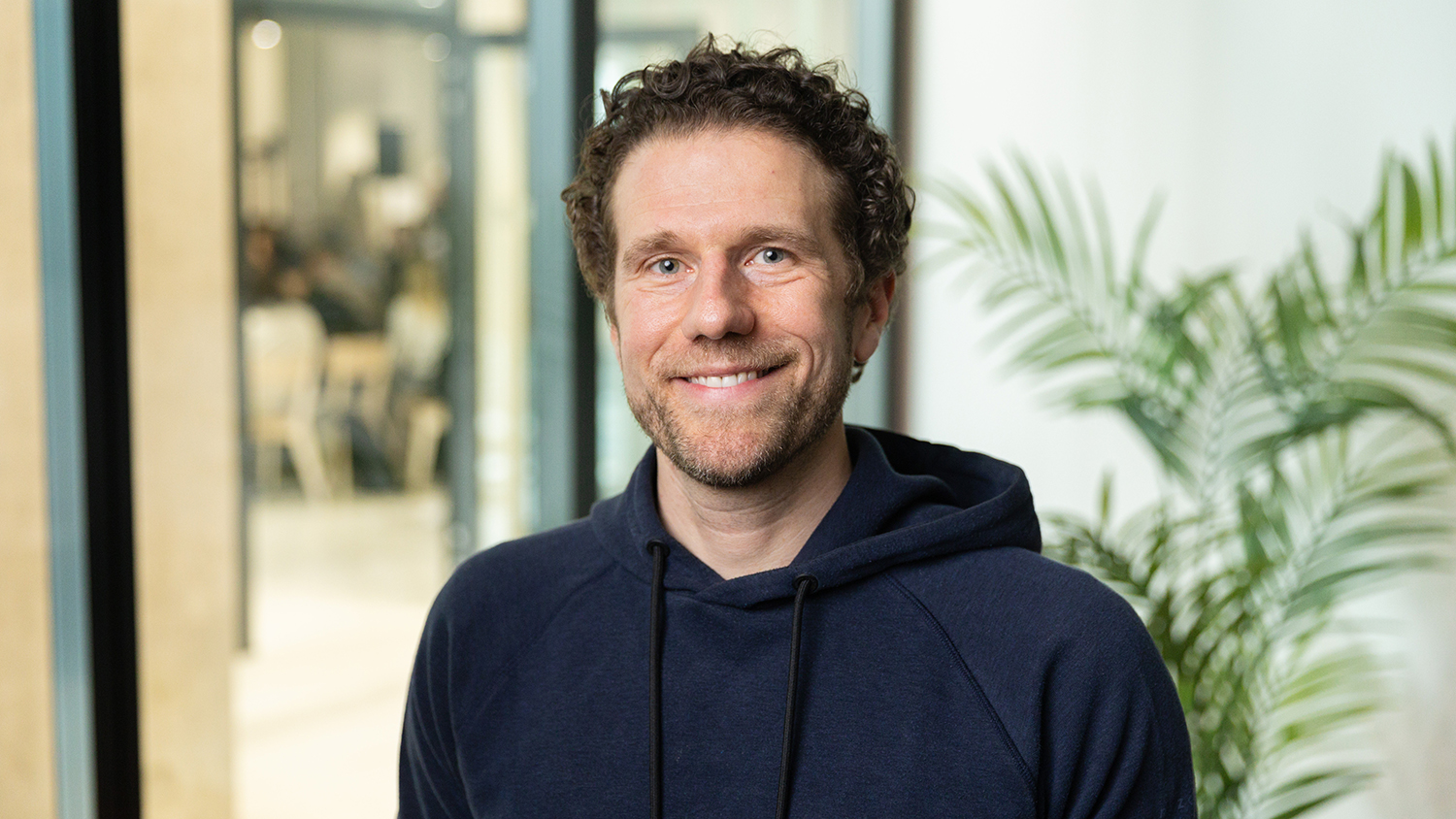 Smiling man with curly hair in a navy hoodie stands indoors near a plant, with a blurred modern office space in the background.
