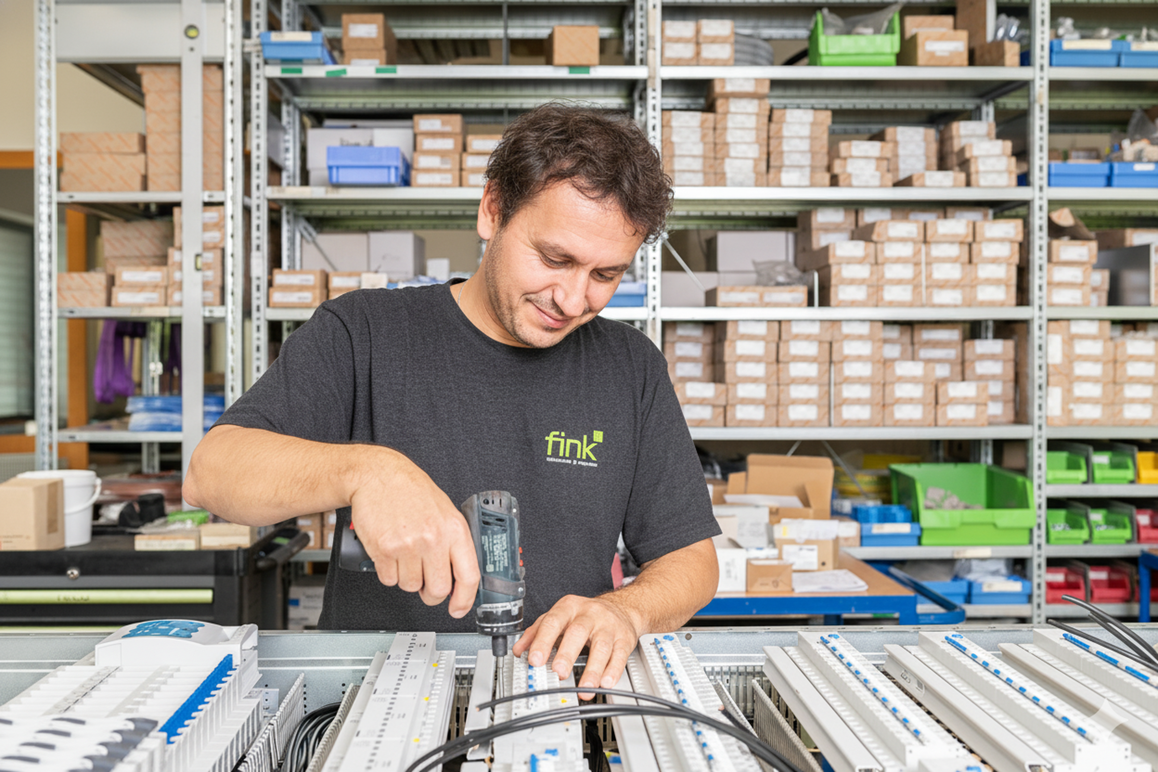 Mann in einer Werkstatt, der mit einer Bohrmaschine an elektrischen Bauteilen arbeitet, umgeben von Regalen mit Kisten und Werkzeugen.