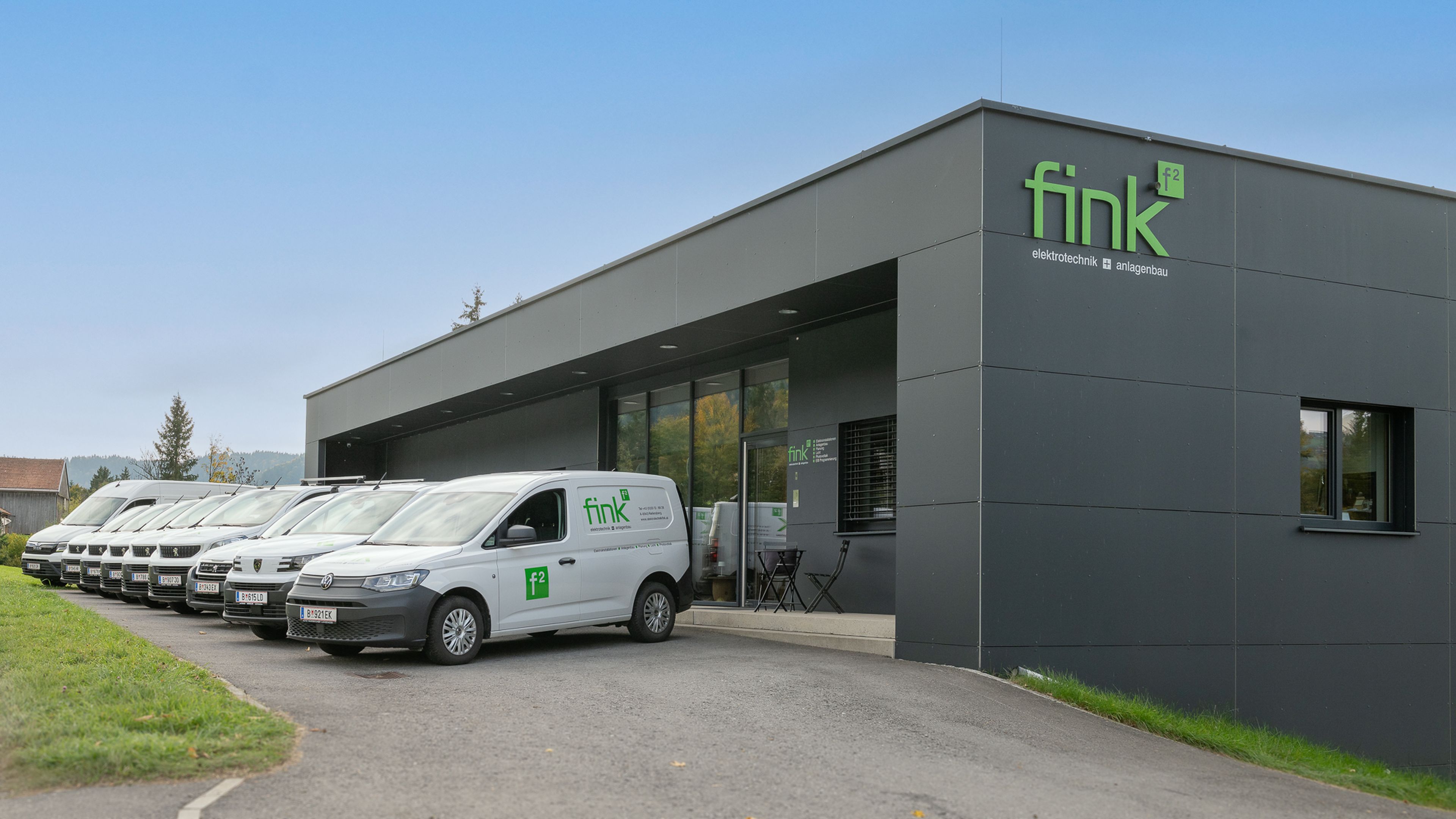 Modernes Gebäude mit „Fink“-Logo, umgeben von mehreren weißen Firmenwagen, die auf einem gepflasterten Bereich unter strahlend blauem Himmel geparkt sind.