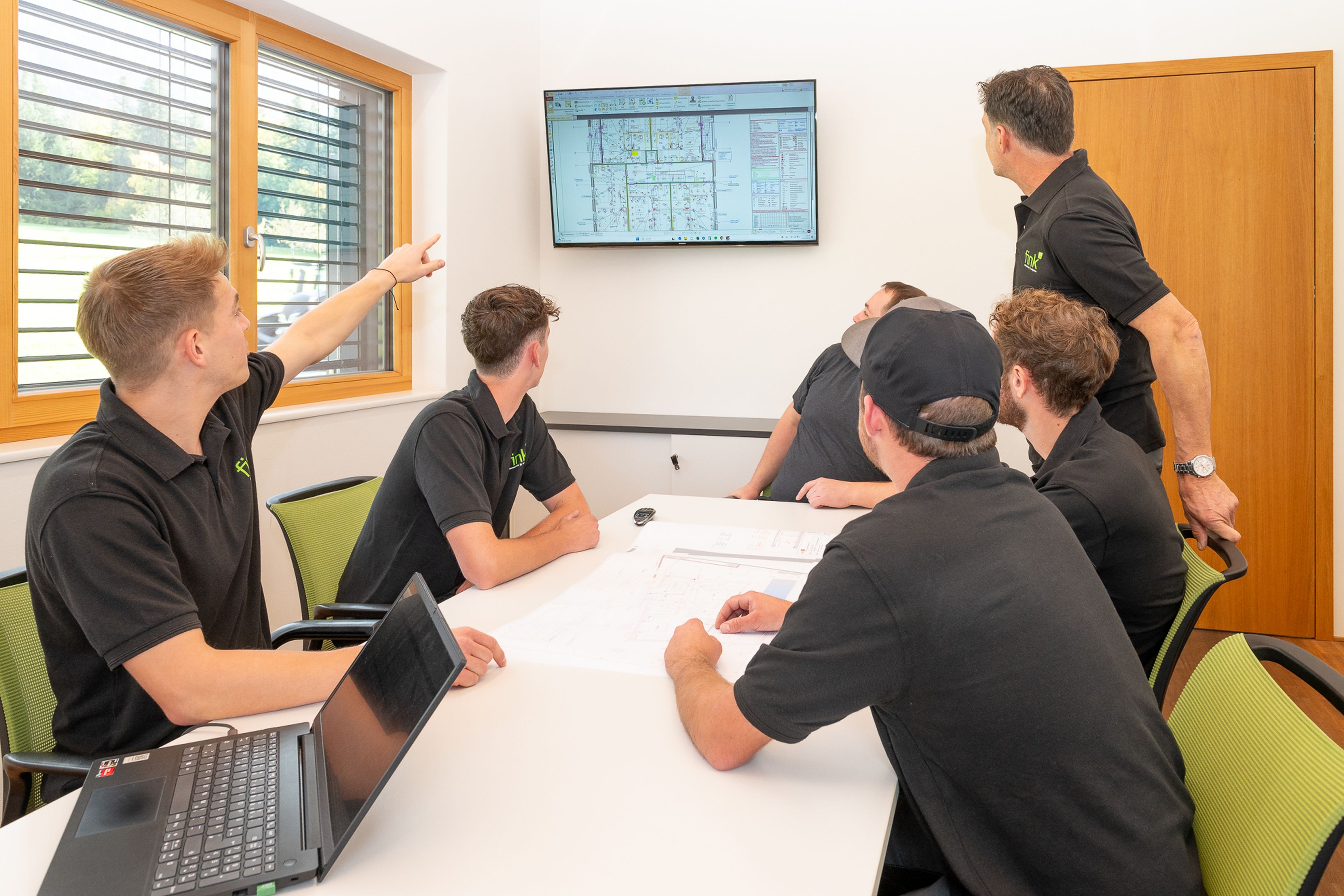 Fünf Personen in schwarzen Hemden sitzen um einen Tisch mit Papieren herum, während einer auf einen an der Wand montierten Bildschirm zeigt, auf dem technische Zeichnungen zu sehen sind.