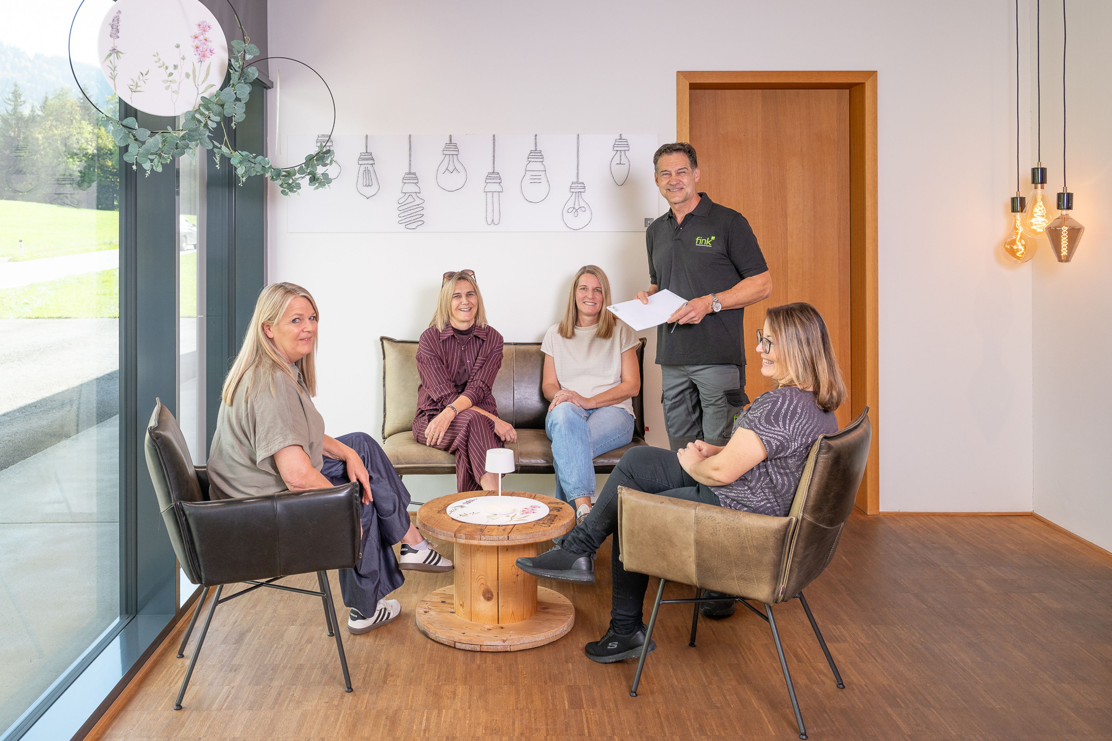 Fünf Personen sitzen und stehen in einem modernen Raum mit Holzboden und unterhalten sich an einem kleinen runden Tisch. Im Hintergrund Wandkunst aus Glühbirnen.