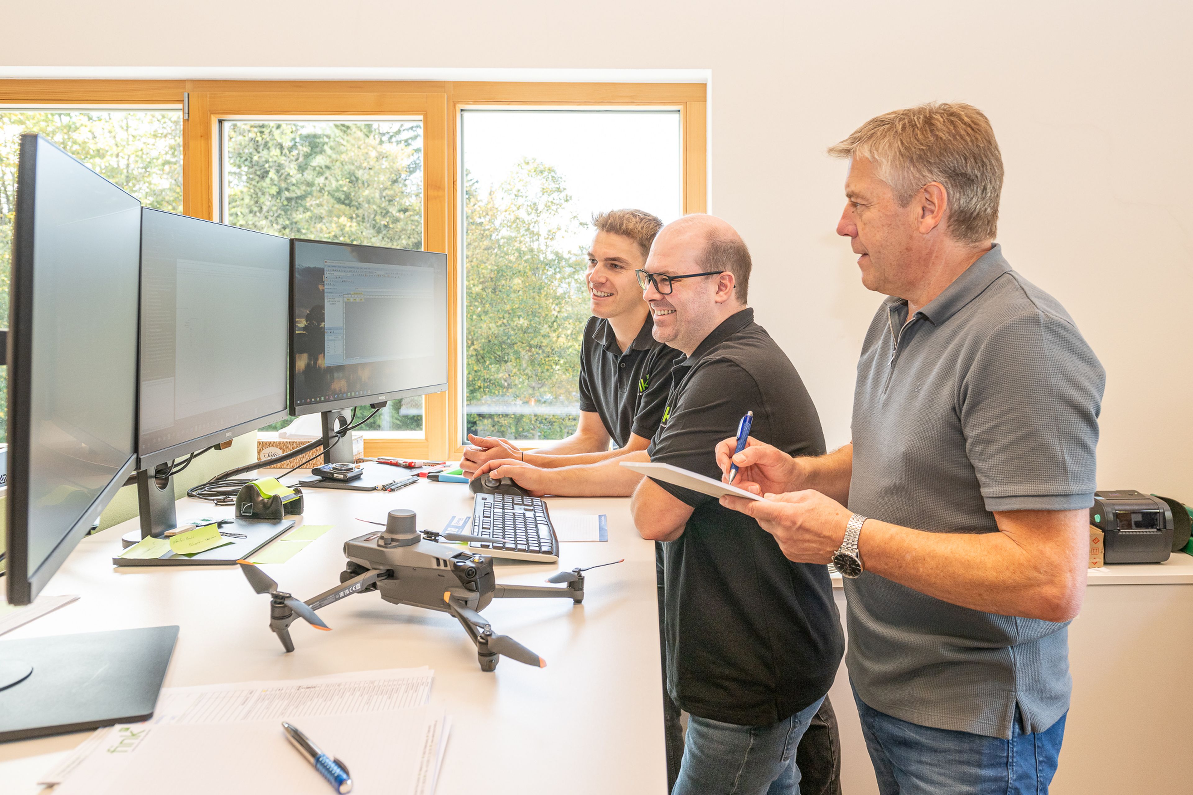 Drei Männer stehen an einem Schreibtisch mit mehreren Monitoren und besprechen ihre Arbeit. Auf dem Schreibtisch liegen eine Drohne und Dokumente. Durch die großen Fenster ist draußen viel Grün zu sehen.