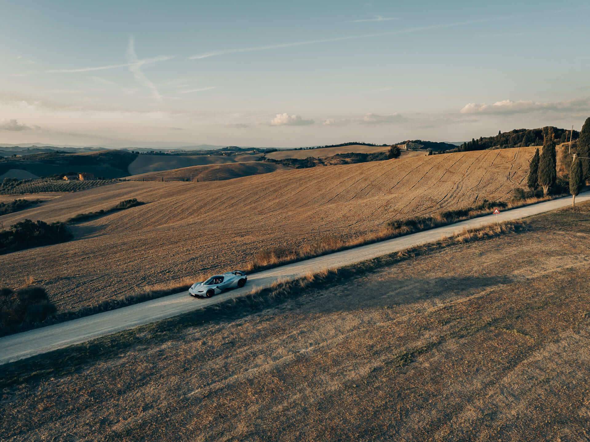 Aerial view of a car driving on a narrow road through vast, rolling fields under a clear sky, with distant hills on the horizon. Aerial view of a car driving on a narrow road through vast, rolling fields under a clear sky, with distant hills on the horizon.