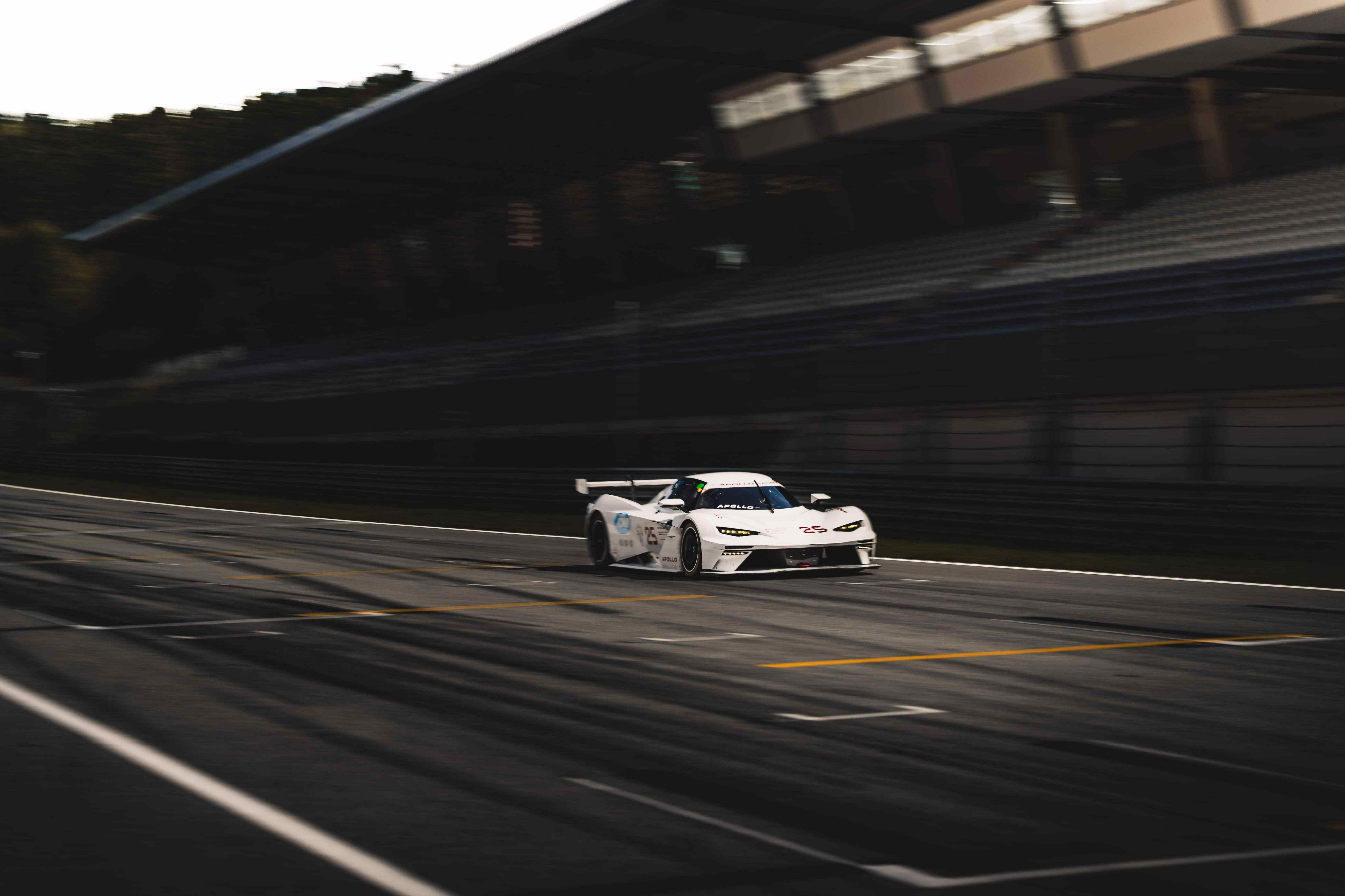 A white race car speeds down an empty track, with blurred grandstands in the background, under a cloudy sky. A white race car speeds down an empty track, with blurred grandstands in the background, under a cloudy sky.